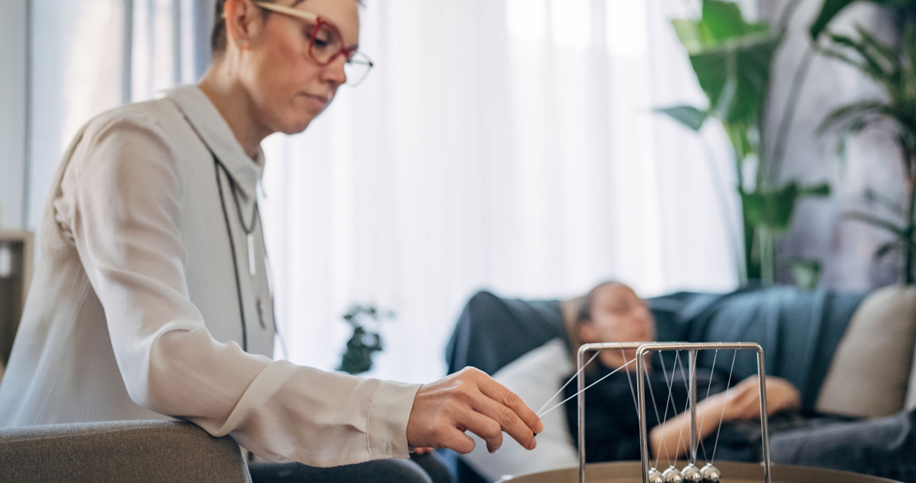 woman-adjusting-Newton's-cradle-on-table-with-hypnosis-client